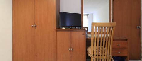Un dormitorio con un armario de madera, una cama con cabecera de madera, una mesita de noche pequeña de madera y un televisor sobre una estantería.