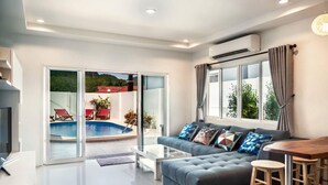 Una sala de estar moderna con un sofá gris, una mesa de centro y un televisor. Hay una puerta corrediza de vidrio que da a una zona de piscina con sillas de descanso rojas.