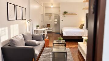 Una sala moderna con un sofá, una mesa de centro y una cama. Hay un comedor con una mesa y sillas, y una pequeña cocina con un refrigerador y una planta.