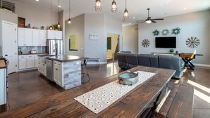 A modern kitchen with white cabinets, a stainless steel refrigerator, and a wooden dining table with a placemat. There is a gray sofa and a ceiling fan in the living area. - Charlotte: Large Groups + Private Hot Tub + Roof Top Deck (Hurricane)