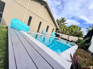 Een zwembad met een houten terras, omgeven door een huis en palmbomen.