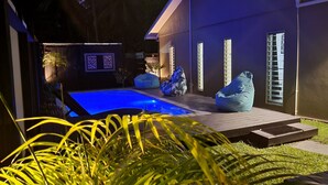 Un espace piscine extérieur moderne, doté d’une piscine illuminée de lumière bleue, d’une terrasse en bois et de deux fauteuils en forme de sac.