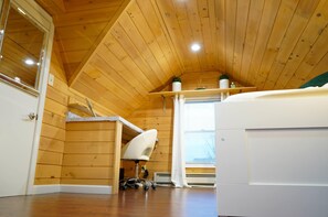Een gezellig kamer in chaletstijl met houten wanden en een schuine kap. Er is een wit bureau met stoel, een raam met zonwering en een klein plankje met decoratieve spullen.