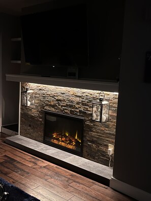 A modern fireplace with a stone facade, a flat-screen TV above it, and a built-in bench with a side table. - Stylish, entire 3-bedroom, 1.5 bath, single house. (Breinigsville)