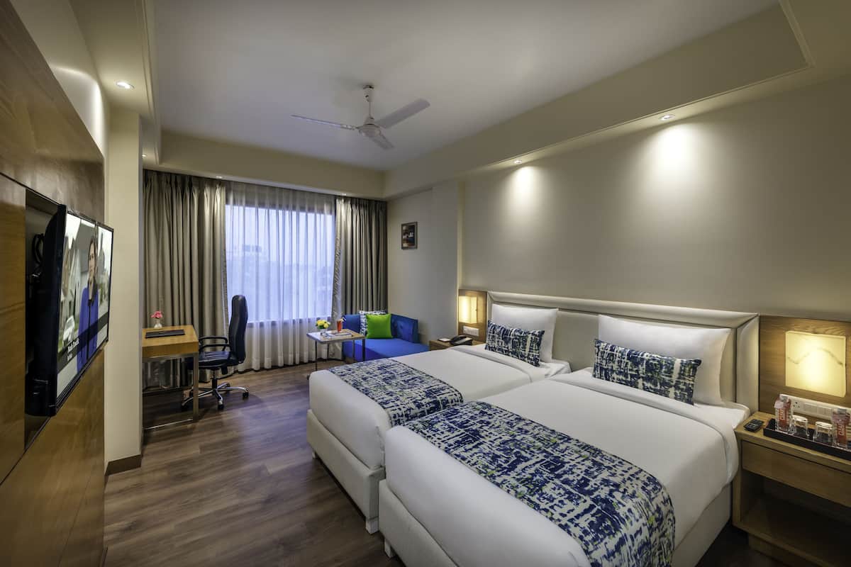 A hotel room with two beds, a desk, a chair, a TV, and a window with curtains.