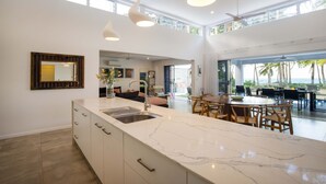 A modern kitchen with a marble countertop, a built-in sink, and pendant lighting. There is a dining area with wooden furniture and a view of the ocean.