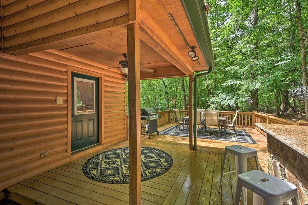 Une cabane en bois dotée d’un porche, d’un coin repas avec des chaises et une table, et offrant une vue sur la forêt environnante.