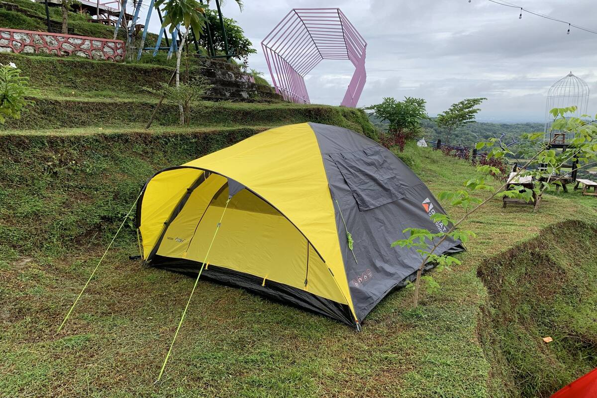 A yellow and gray tent set up on a grassy slope with a red and white structure in the background.