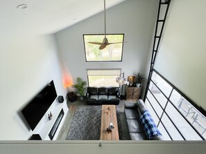 Een moderne woonkamer met een zwarte leren bank, een houten salontafel en een televisie links. Twee ramen zorgen voor natuurlijk licht en uitzicht op de omgeving.
