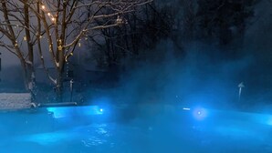 Zona de piscina exterior climatizada con niebla e iluminación azul, rodeada de árboles y un paisaje nevado.