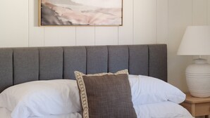 Un lit bien fait, avec une tête de lit grise, des oreillers blancs et à motifs, et un grand tableau abstrait accroché au mur au-dessus.