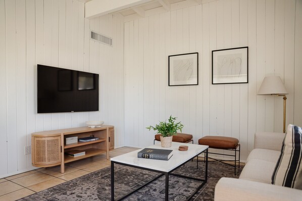 Ein modernes Wohnzimmer mit einem wandmontierten Flachbildfernseher, einem weißen Couchtisch, einem braunen Lederhocker und einem beigen Sofa.