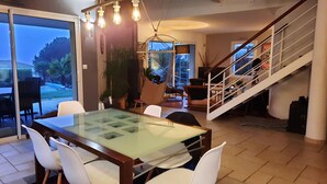 A dining area with a glass-top table and white chairs, a hanging light fixture, and a staircase leading to an upper level.