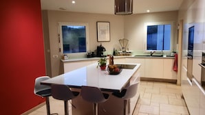 A modern kitchen with a central island, bar stools, a coffee machine, and a potted plant.