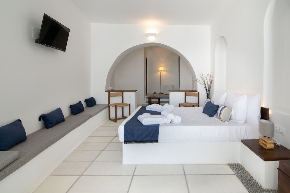 Ein modernes Schlafzimmer mit einem großen Bett, einem an der Wand montierten Fernseher, einer Sitzecke mit Kissen und einem kleinen Tisch mit einer Lampe.