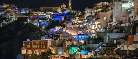 A densely packed hillside town with buildings illuminated at night, featuring a prominent clock tower and a variety of architectural styles.