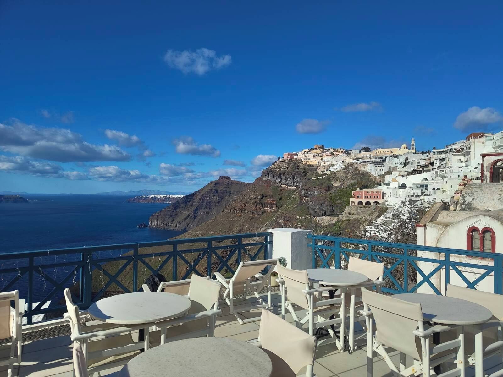 Eine Terrasse am Meer mit weißen Tischen und Stühlen, die auf eine Küstenstadt mit weißen Häusern und Blick aufs Meer blickt.