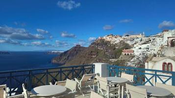 A seaside terrace with white tables and chairs, overlooking a coastal town with white buildings and a view of the sea.
