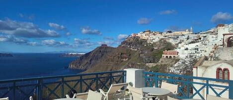 A seaside terrace with white tables and chairs, overlooking a coastal town with white buildings and a view of the sea.