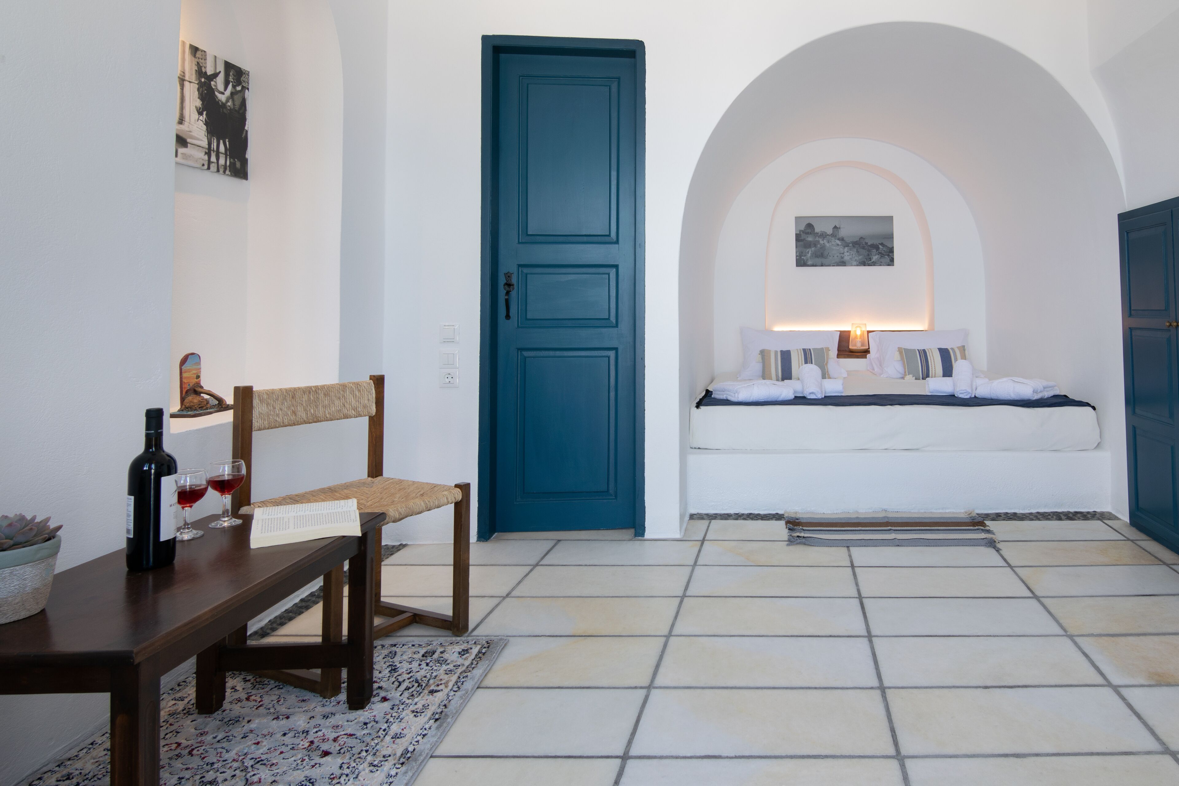 A room with a wooden bench, a table with a bottle and glasses, a blue door, a bed with pillows, and a wall-mounted lamp.