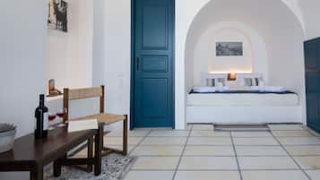 Een kamer met een houten bankje, een tafel met een fles en glazen, een blauwe deur, een bed met kussens en een wandlamp.