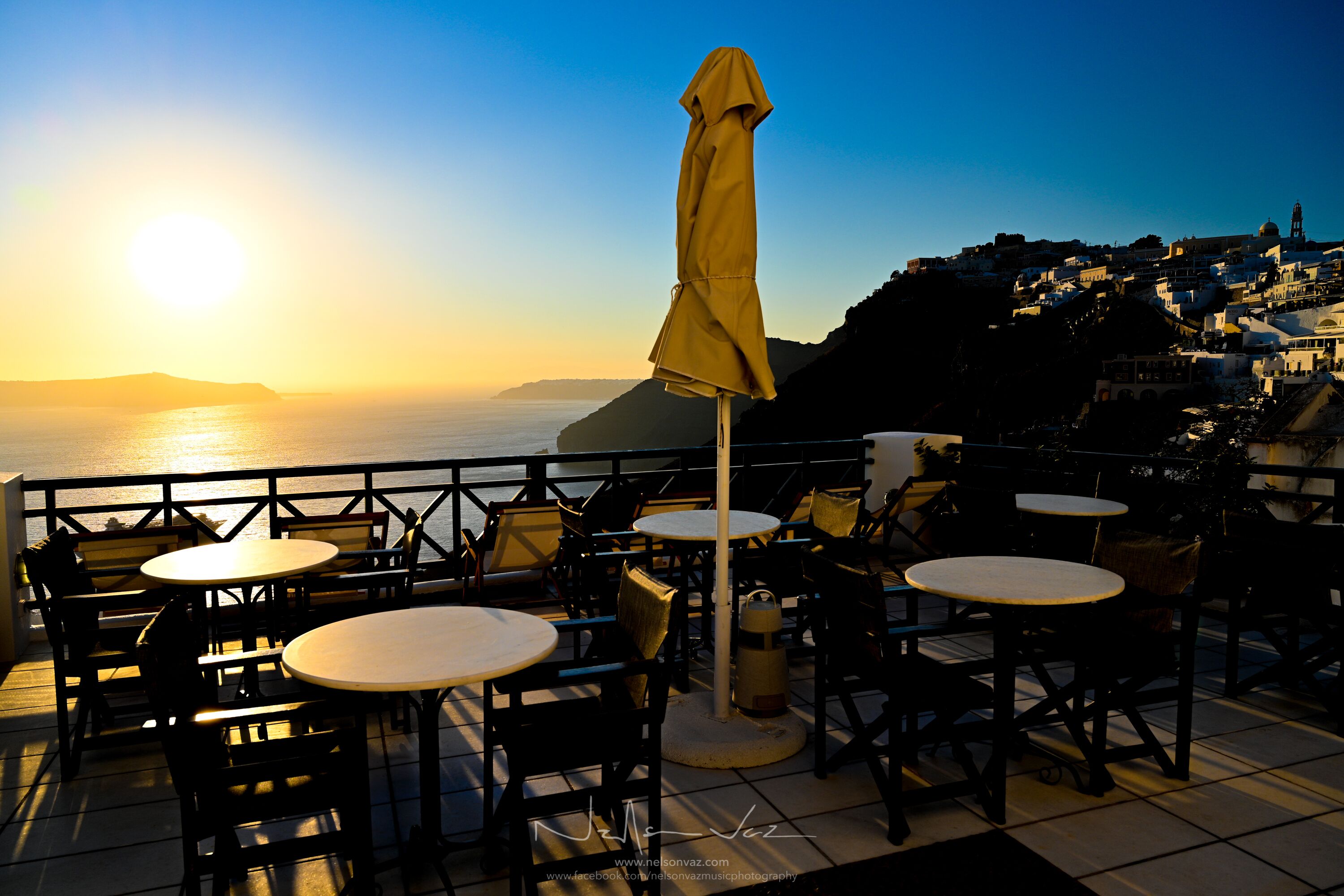 Una terraza con mesas y sillas redondas, un paraguas amarillo y vistas al mar y a un pueblo en la distancia.