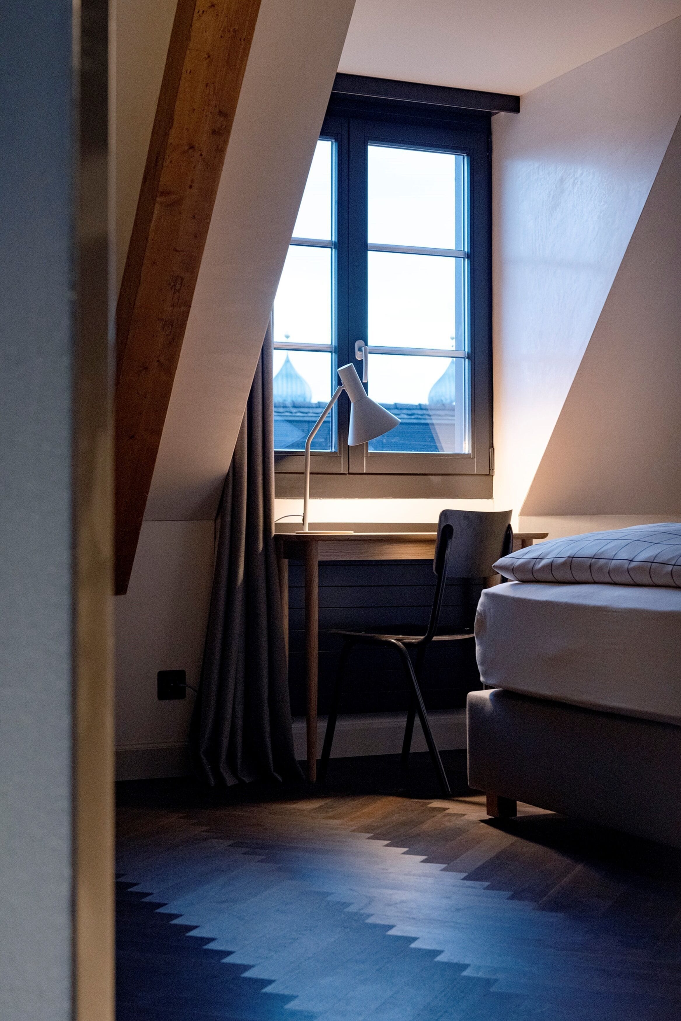 A bedroom with a sloped ceiling, a desk with a chair, a bed, and a window with curtains.