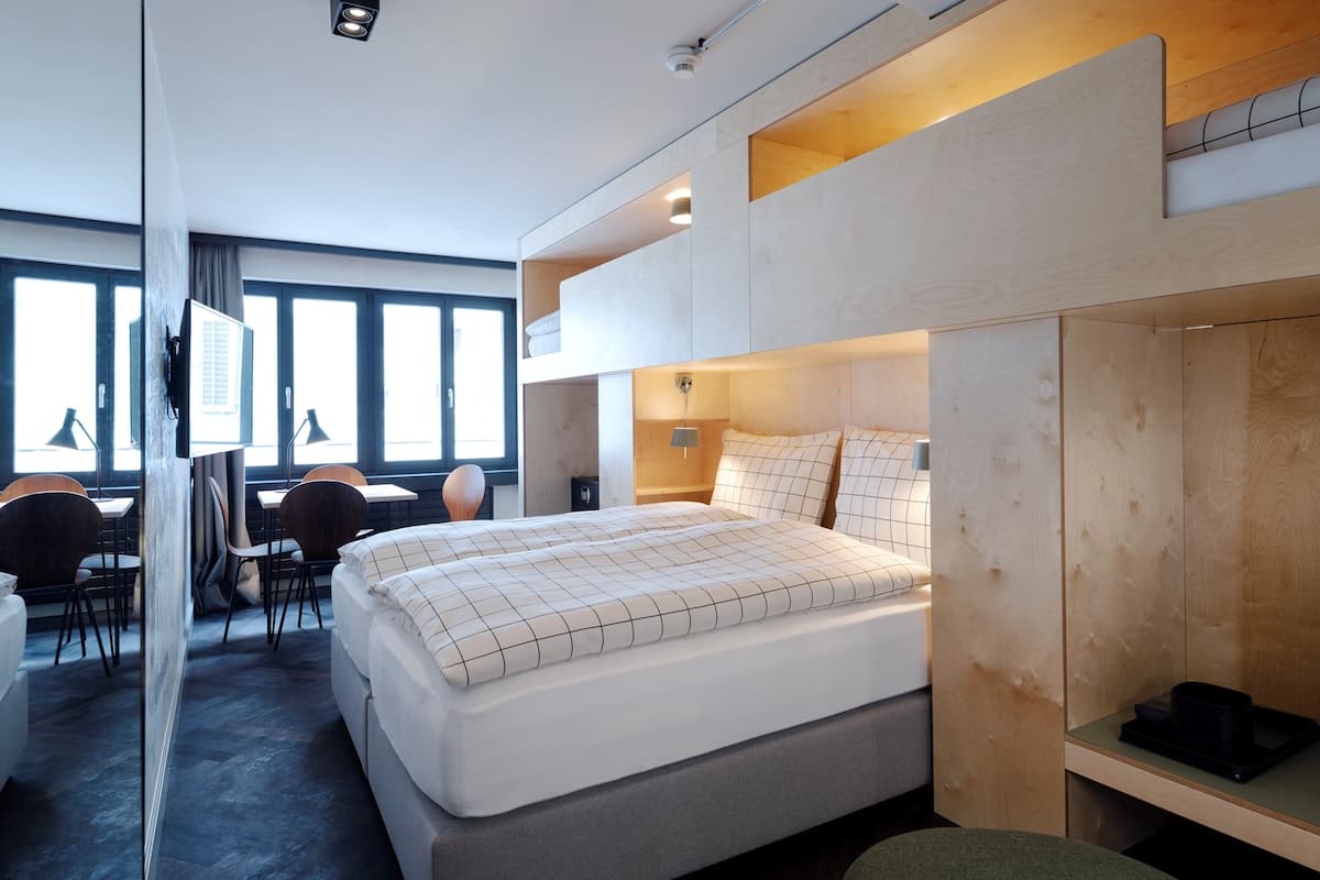 A modern hotel room with a large bed, a desk with chairs, and a view through large windows.