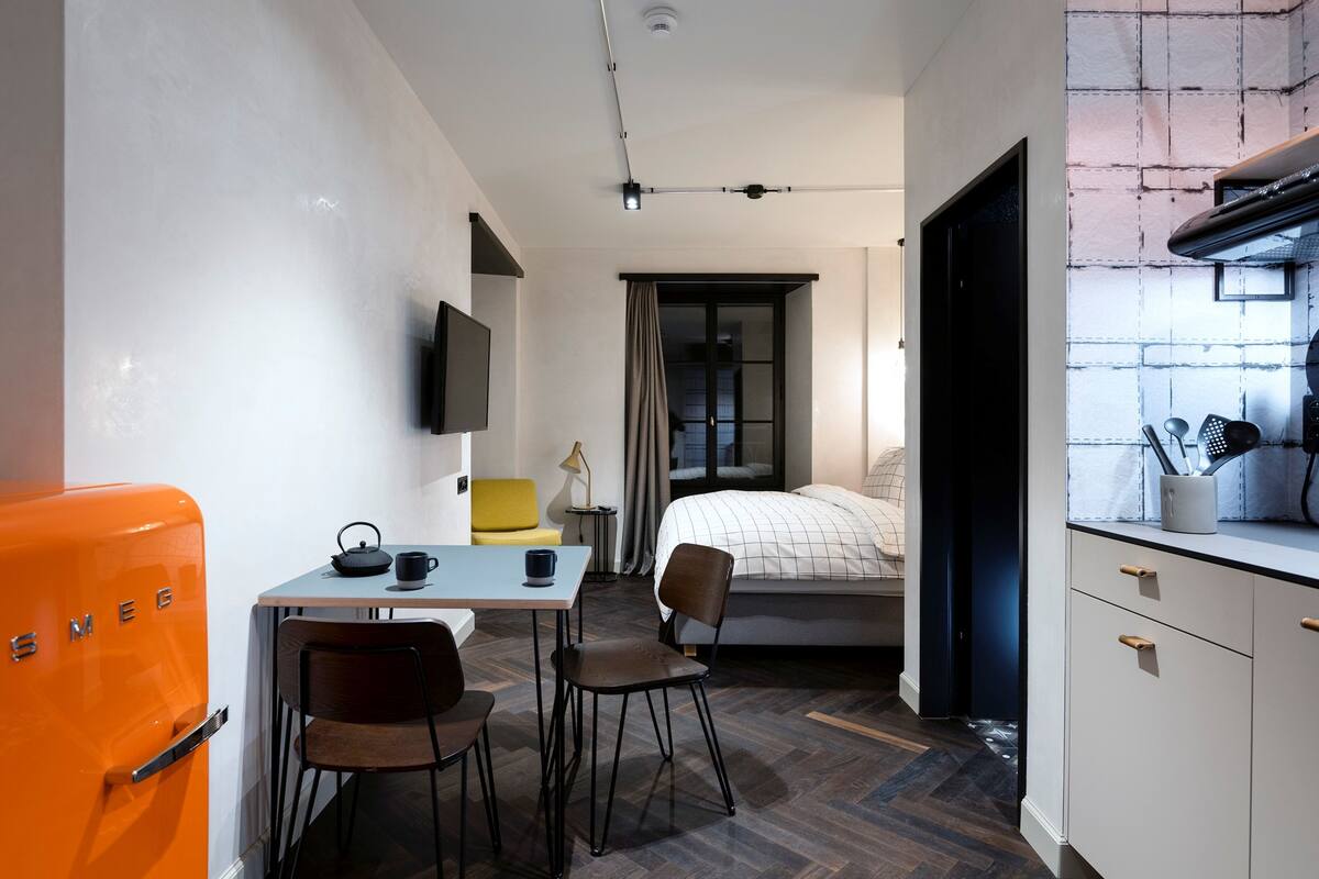 A compact hotel room with a bed, a small table with chairs, a kitchenette, and a television on the wall.