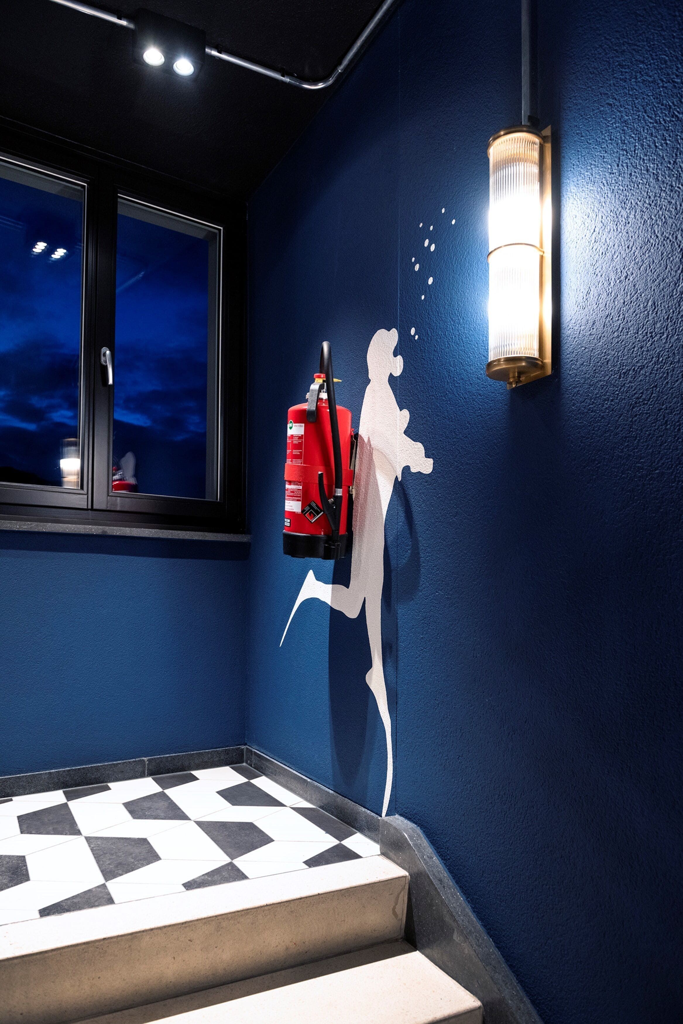 Quarto com parede azul, janela com vista do céu noturno, extintor de incêndio fixado na parede e piso estampado.