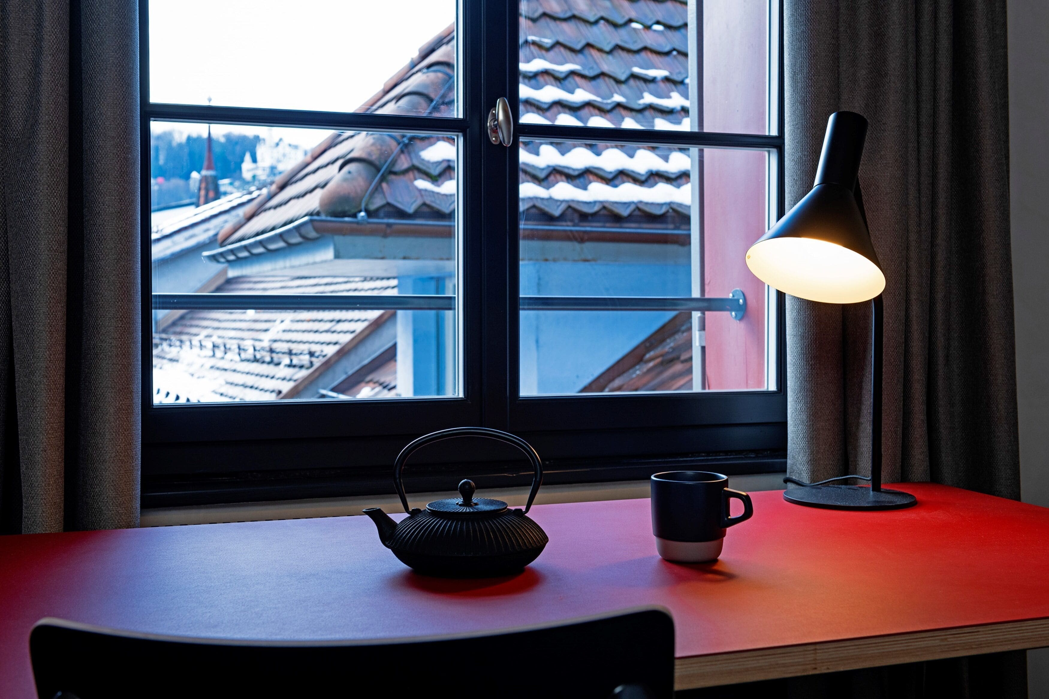 Une table avec une théière et une tasse, une lampe et une fenêtre donnant sur des bâtiments.