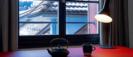 Une table avec une théière et une tasse, une lampe et une fenêtre donnant sur des bâtiments.