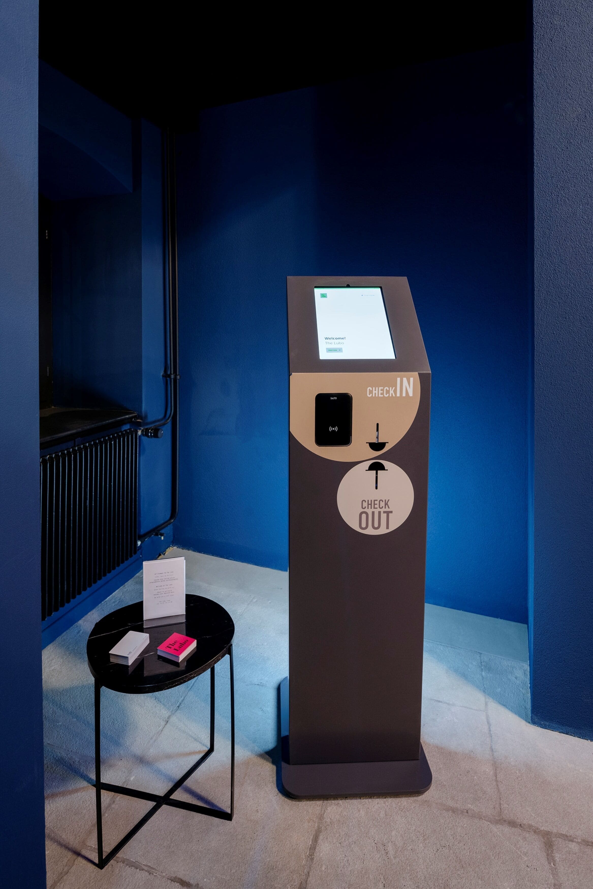 Un quiosco de registro de entrada y salida con pantalla digital, situado junto a un radiador y una pequeña mesa con un libro y un bloc de notas.