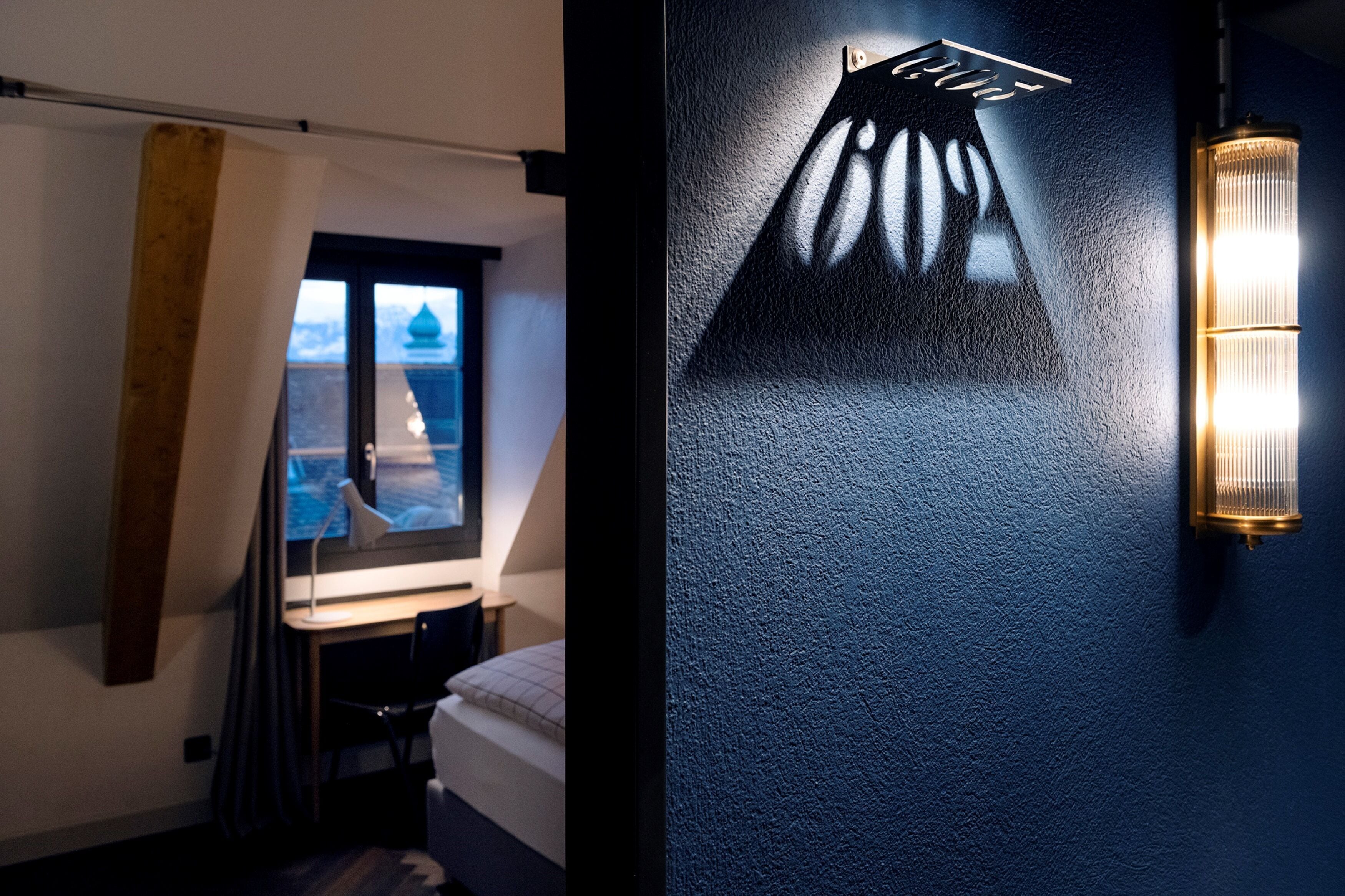 A room with a bed, a desk, and a window. There is a wall-mounted light fixture and a curtain.