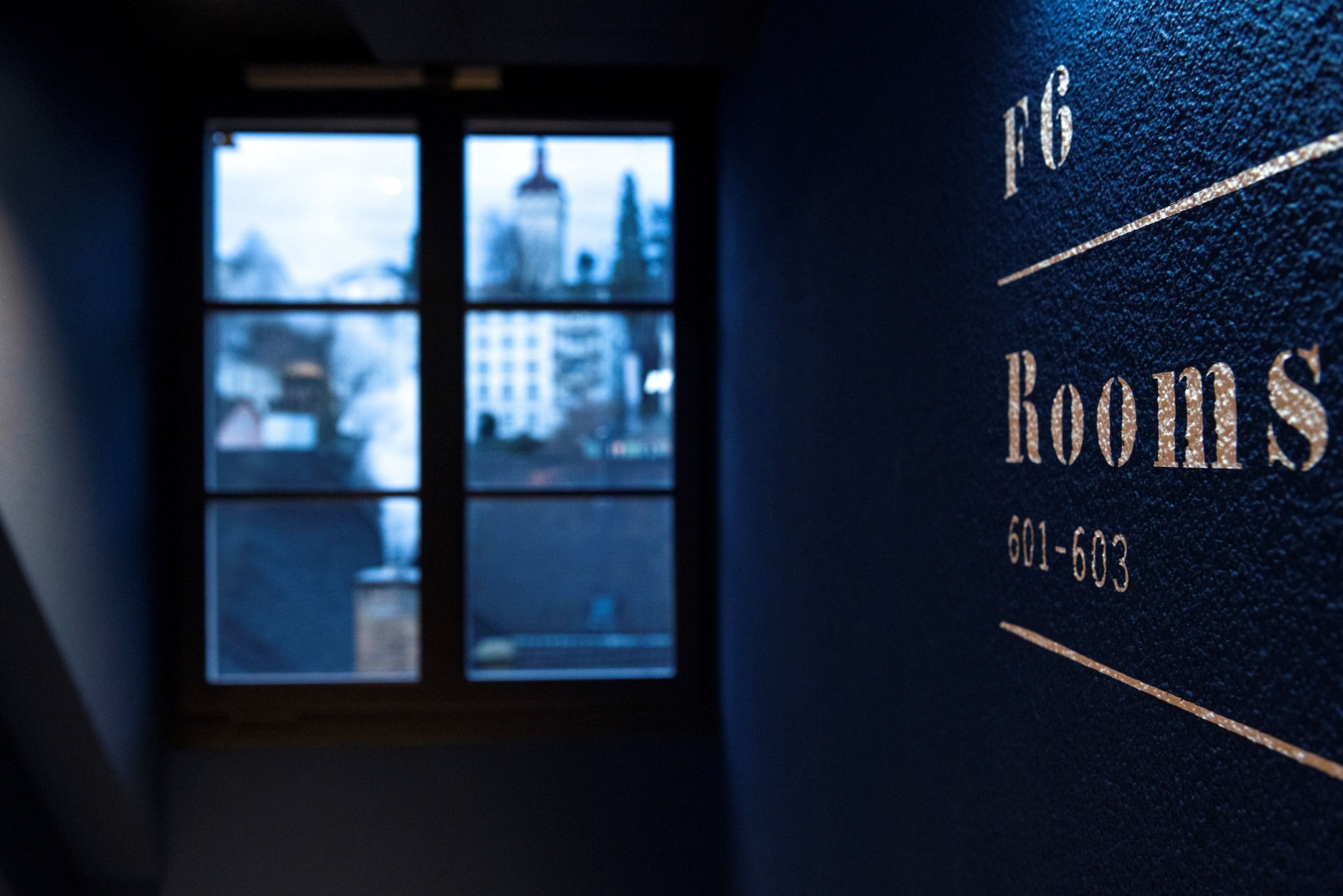 Ein dunkler Raum mit einem Fenster, das einen Stadtblick zeigt. Der Schriftzug '16 Rooms' und die Telefonnummer '601-603' sind sichtbar.