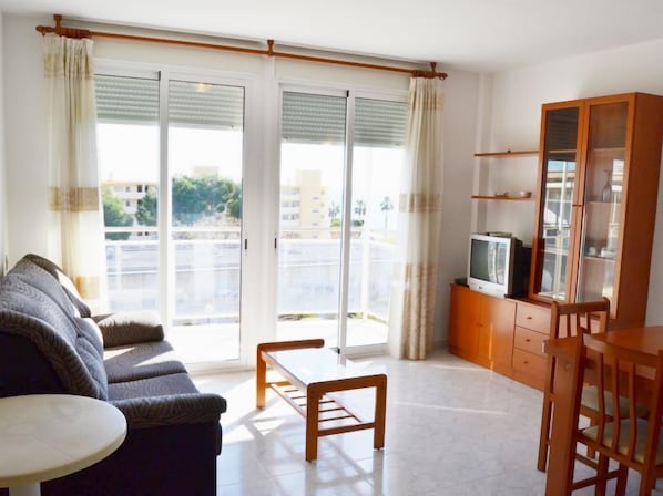 A living room with a sofa, a coffee table, a TV on a wooden cabinet, and a sliding glass door leading to a balcony.