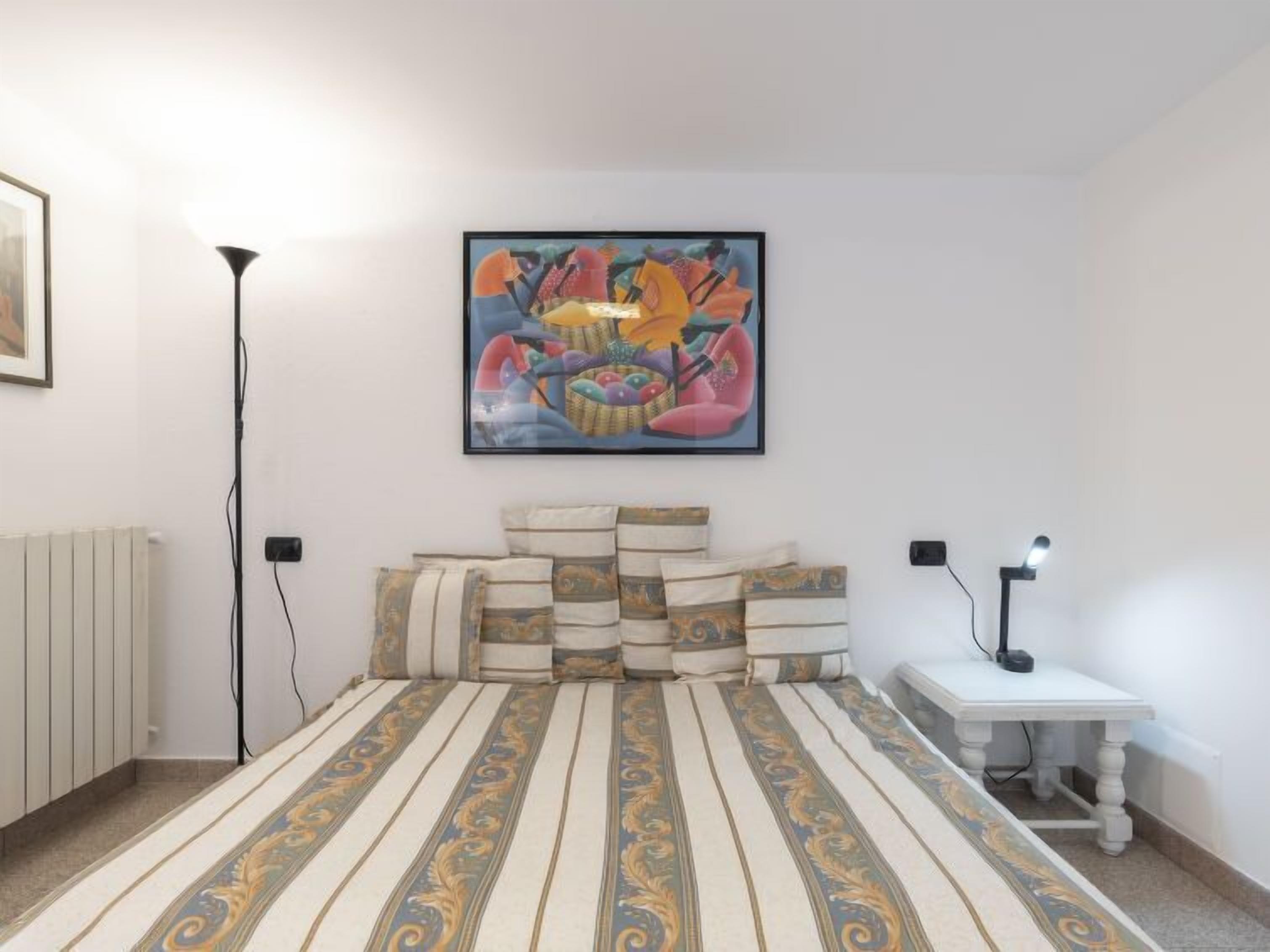 A bedroom with a large bed, a bedside table, a floor lamp, and a painting on the wall.