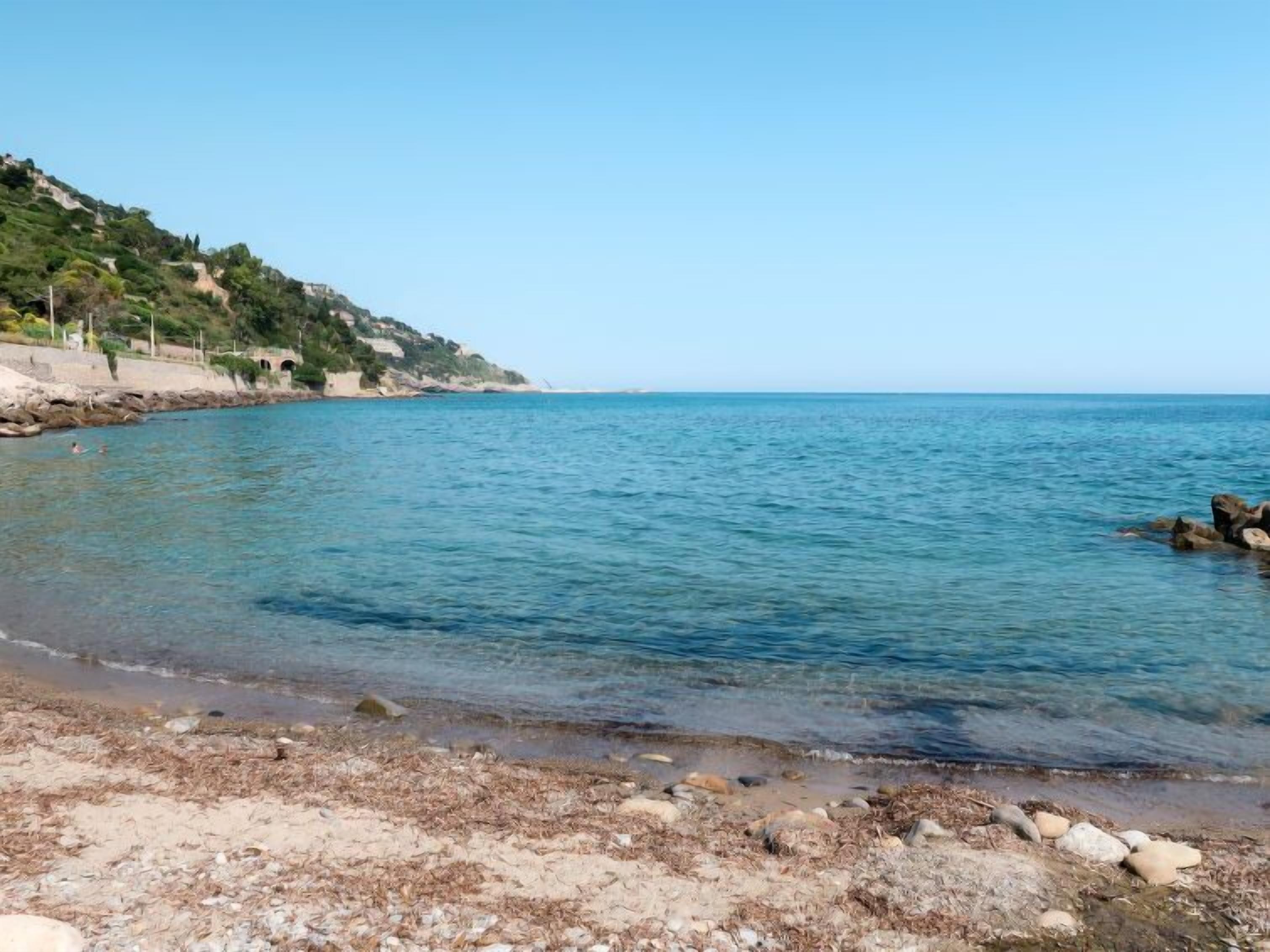 Une plage à l’eau d’un bleu limpide, avec un rivage rocheux et une colline couverte de végétation.