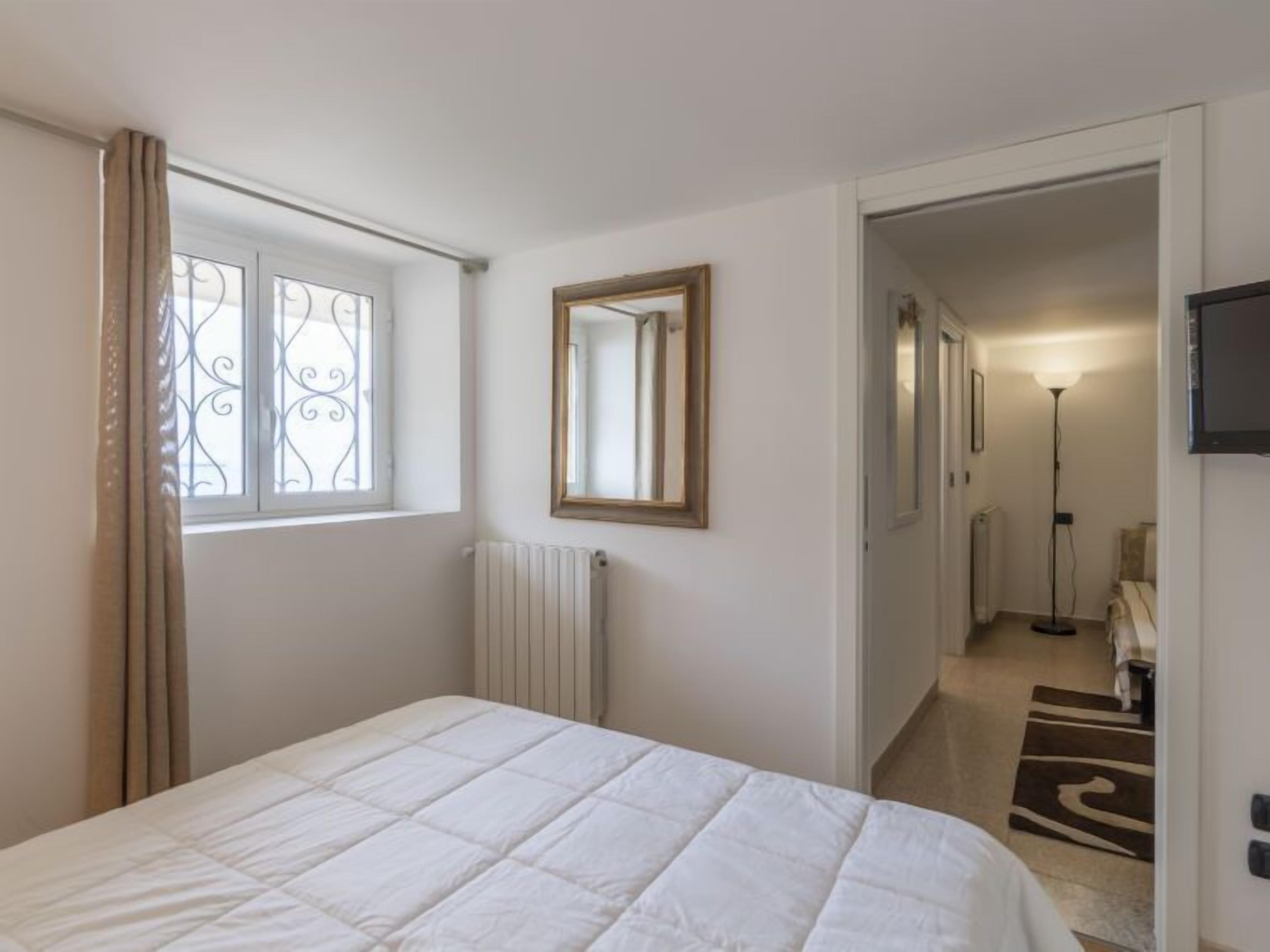 Une chambre à coucher avec un lit, une fenêtre avec des rideaux, un radiateur, un miroir et un couloir menant à une autre pièce.