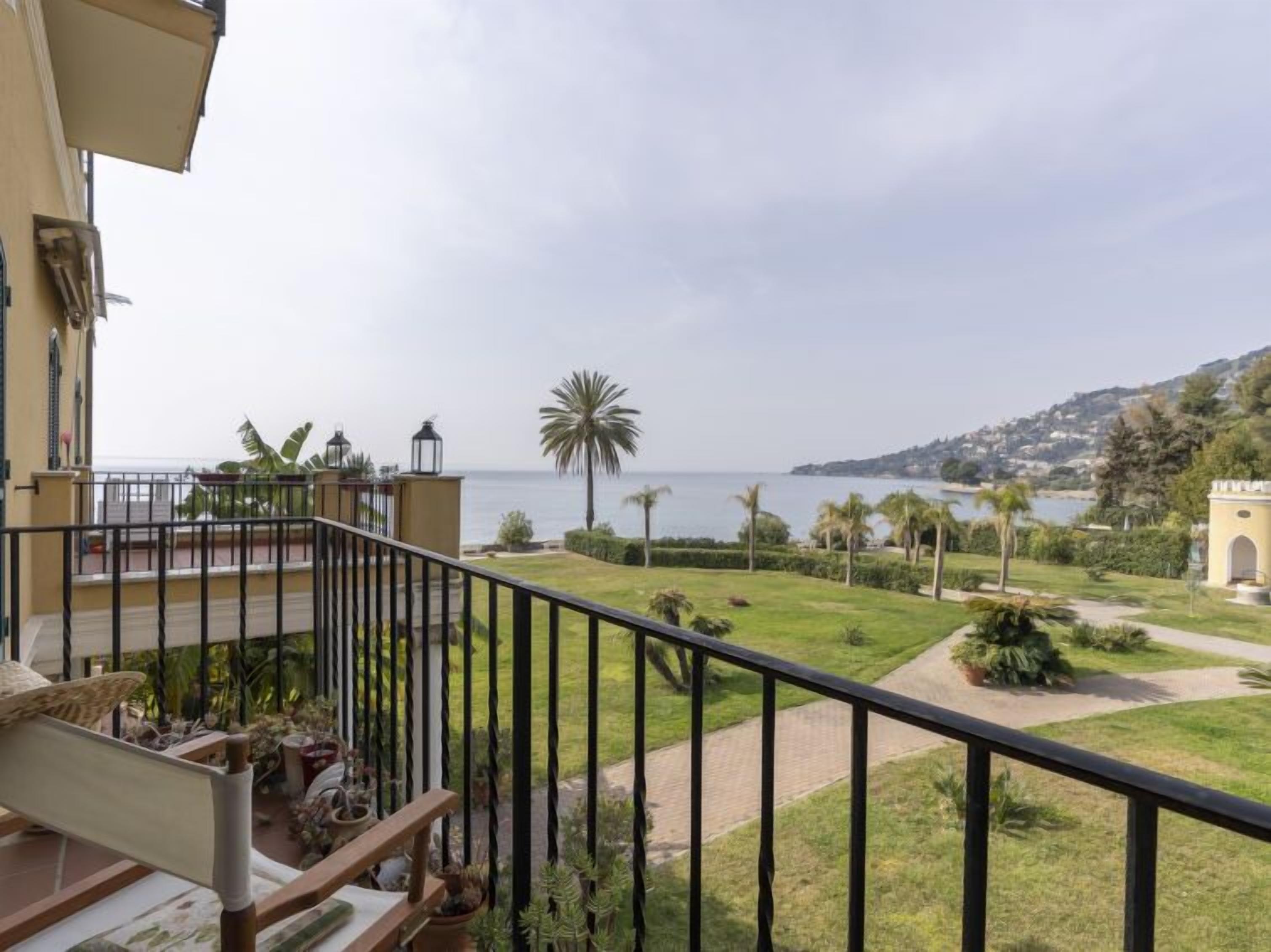 A balcony with a view of the sea, a palm tree, and a pathway leading to a building with an archway.