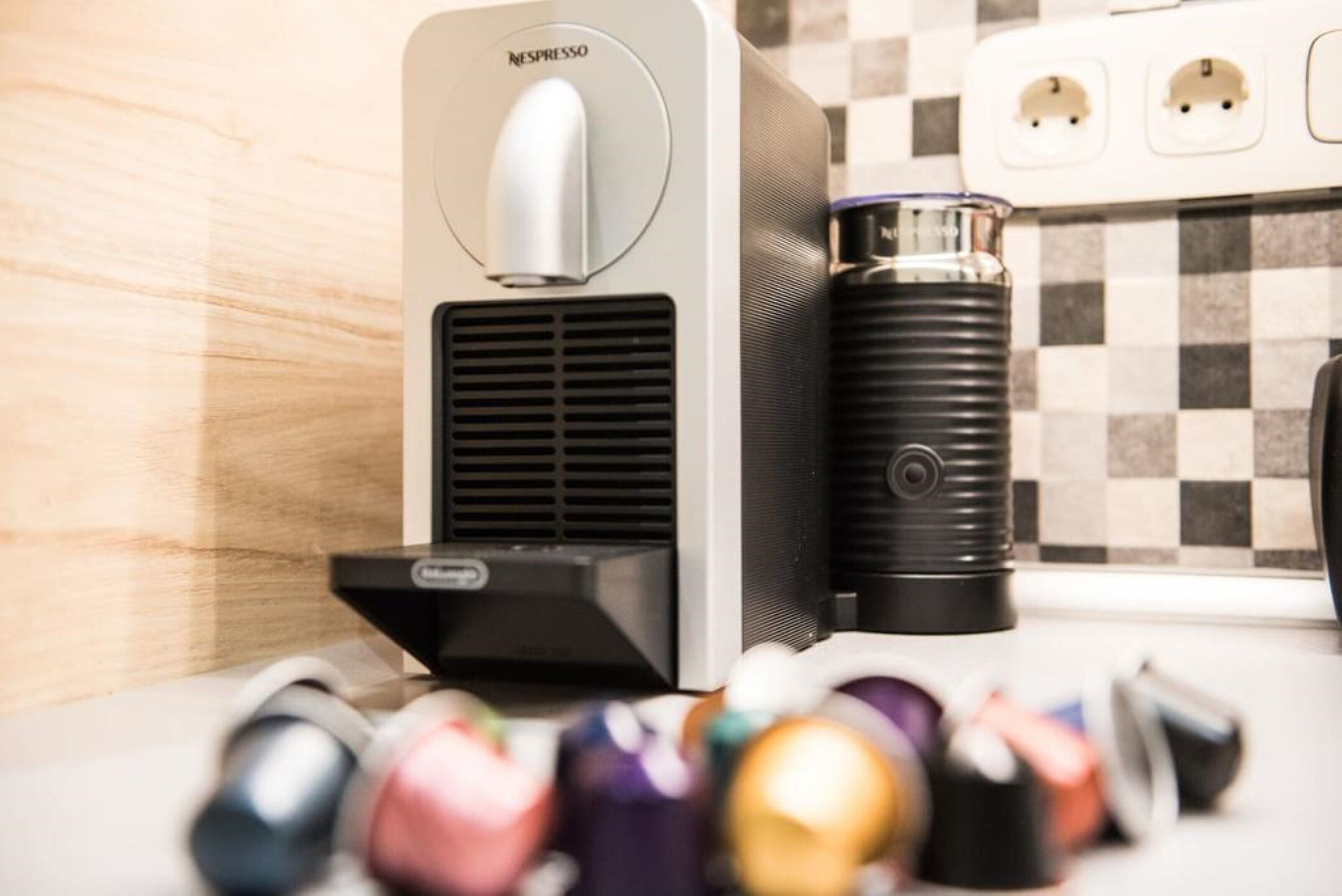 Une machine à café Nespresso et un distributeur d’eau sont posés sur un plan de travail de cuisine, avec un pare-éclaboussures à carreaux.