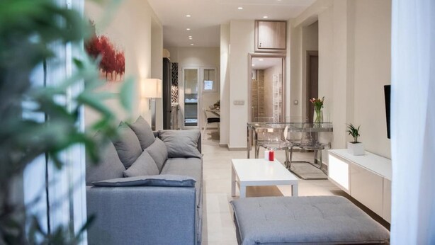 A modern living room with a sofa, a coffee table, and a TV. There is a kitchen area with a dining table and chairs.