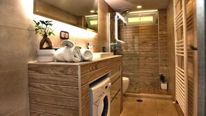 A modern bathroom with a wooden vanity, a built-in washing machine, a glass shower enclosure, and a towel rack.