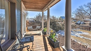 A covered porch with a wooden deck, two chairs, a bench, and planters with yellow flowers. - Historic Old Colorado City Home w/ Backyard! (Colorado Springs)