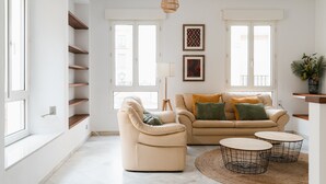 A spacious living room with a beige sofa, a cream armchair, and two round coffee tables. Shelves are built into the wall, and there are two windows providing natural light. - Real Carretería. 3 bedrooms, 3 bathrooms, terrace, views (Sevilla)