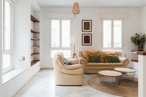 A spacious living room with a beige sofa, a cream armchair, and two round coffee tables. Shelves are built into the wall, and there are two windows providing natural light. - Real Carretería. 3 bedrooms, 3 bathrooms, terrace, views (Sevilla)