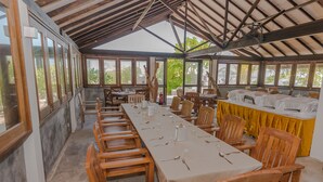 A long dining table set with chairs, a yellow tablecloth, and a fan in a covered outdoor area with large windows. - Premier Beach (Gulhi)