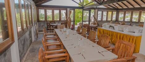 Una larga mesa de comedor dispuesta con sillas, mantel amarillo y un ventilador en un espacio exterior cubierto con ventanas grandes.