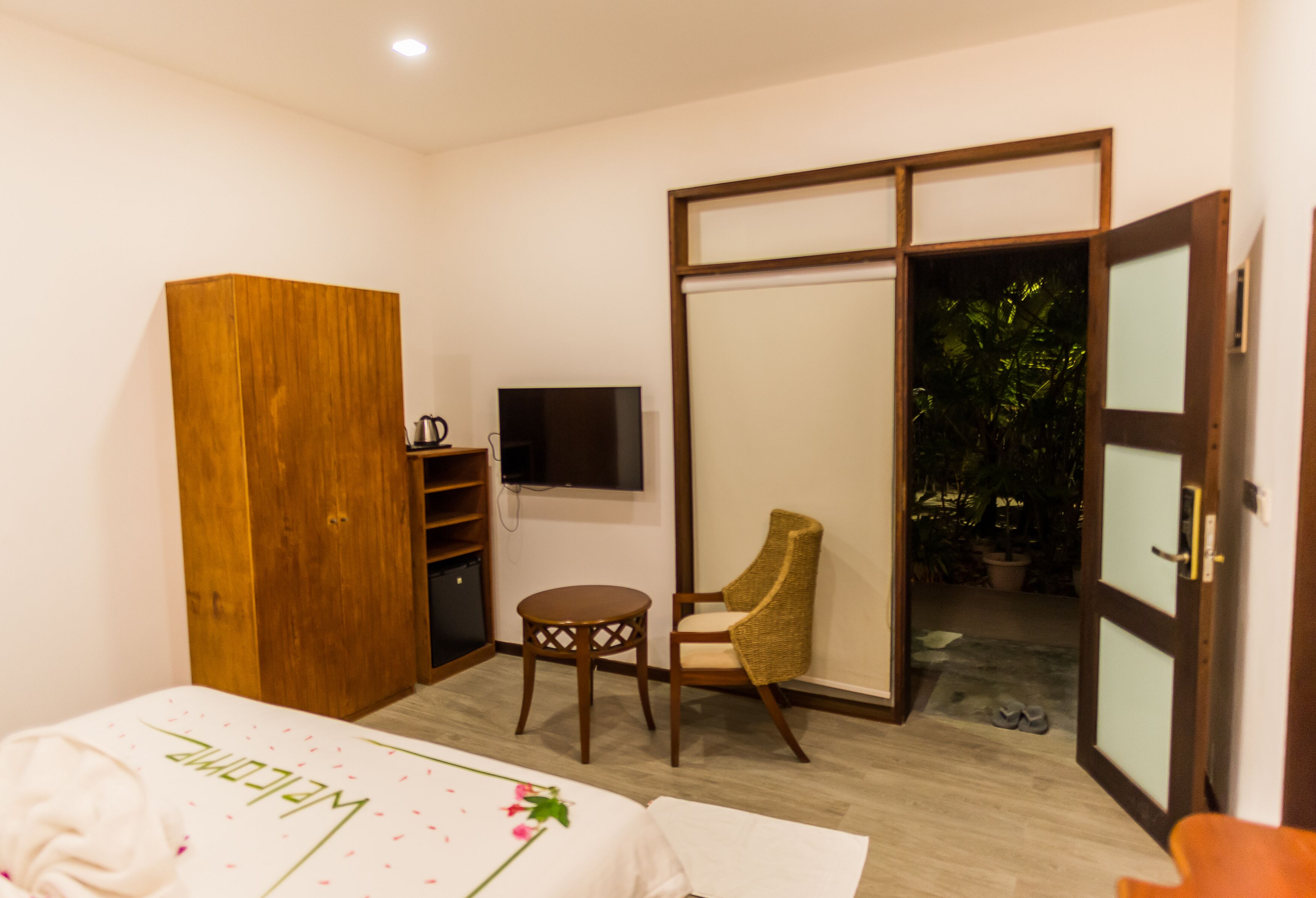 Un dormitorio con cama, armario, televisor, una silla y puerta corredera que da a un balcón con plantas.