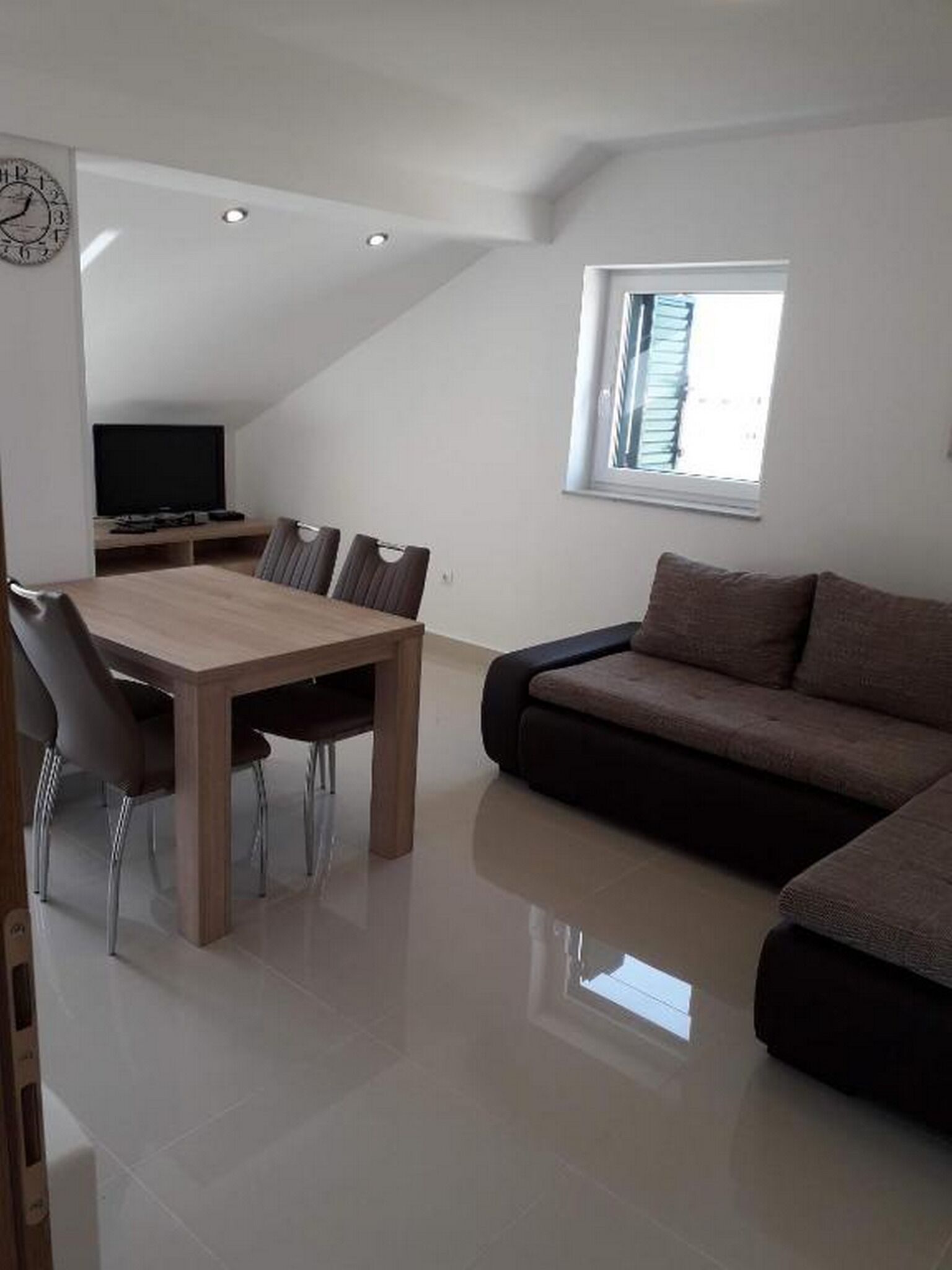 A modern living room with a wooden dining table, chairs, a sofa, a flat-screen TV, and a window with blinds.